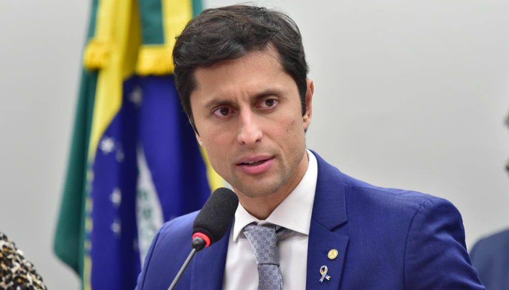Em fotografia, centralizado até a altura do peito, falando em um microfone e olhando na direção lateral à fotografia, aparece Duarte Junior. Ele é um homem branco, cabelos lisos pretos, olhos castanhos, usa um paletó azul, com camiseta branca internamente e gravata cinza. Ao fundo a bandeira do Brasil, ao longe uma parede branca. Fim da descrição.